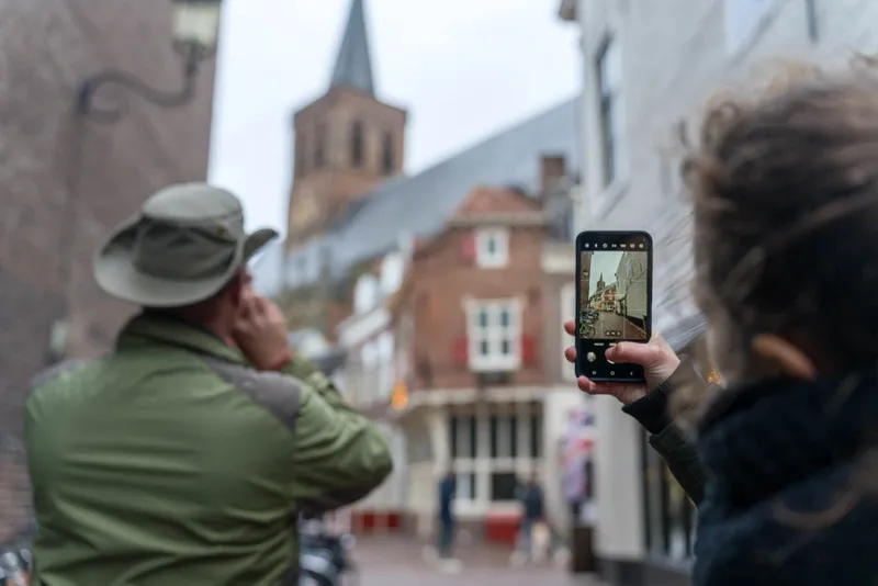 Een deelnemer met een hoed en een deelnemer met krullen fotograferen een kerktoren in een winkelstraat, op het scherm de foto