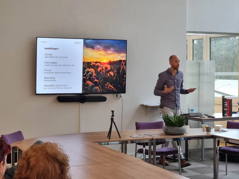 Maarten geeft uitleg bij een scherm met een foto van een zonsondergang, voor een groep aan een lange tafel
