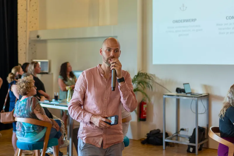 Maarten met een microfoon in de hand voor een zaal met deelnemers, op het scherm de onderwerpen van de workshop