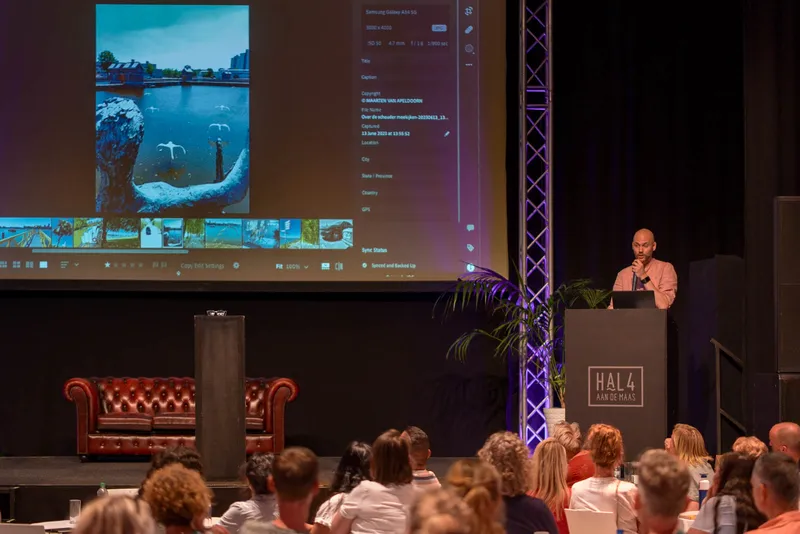 Maarten op een podium voor een groot scherm met een foto van een zwaan, de zaal kijkt toe