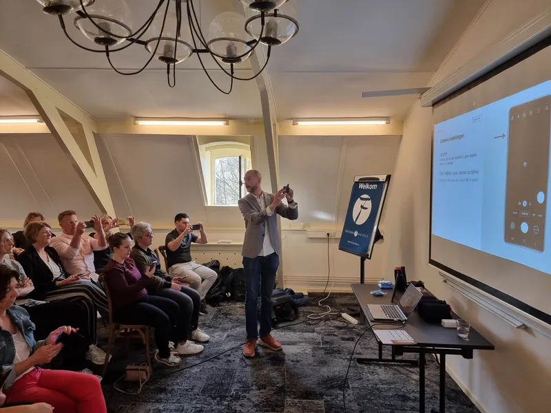 Maarten fotografeert voor een zaal onder een houten kap, de deelnemers houden hun telefoon omhoog, op het scherm de camera-app