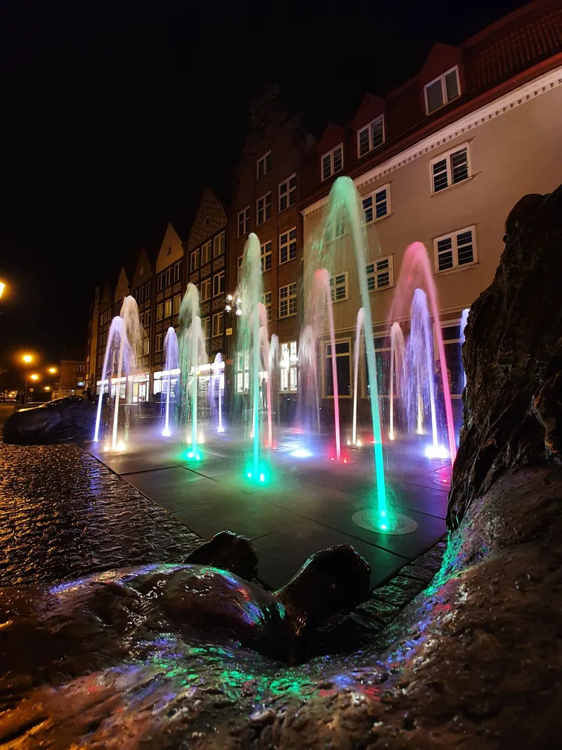 Verlichte fonteinen in roze, groen en blauw bij nacht, met een rij oude gevels erachter