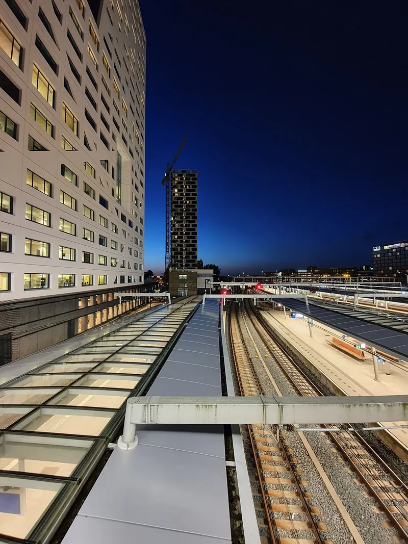 Perrons en sporen van een station bij schemering, met een hoog verlicht gebouw ernaast