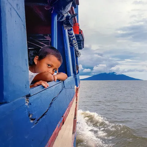 Jongen kijkt uit het raam van een blauwe boot, met een vulkaan aan de horizon